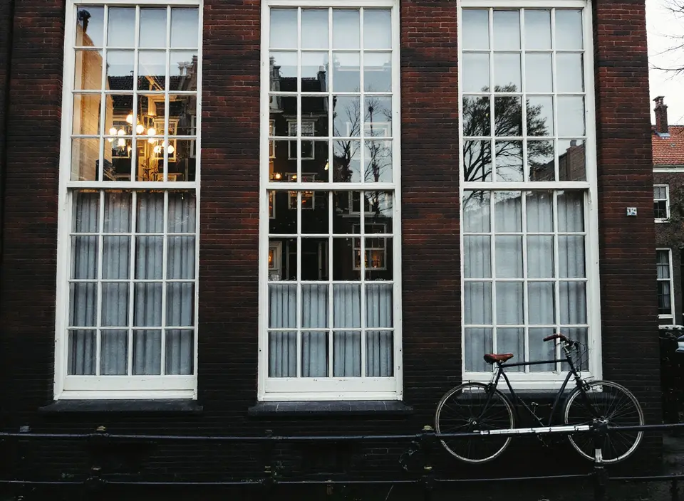 Gevel van een grachtenpand met hoge witte schuiframen met roeden en oud glas, een fiets bij het hek